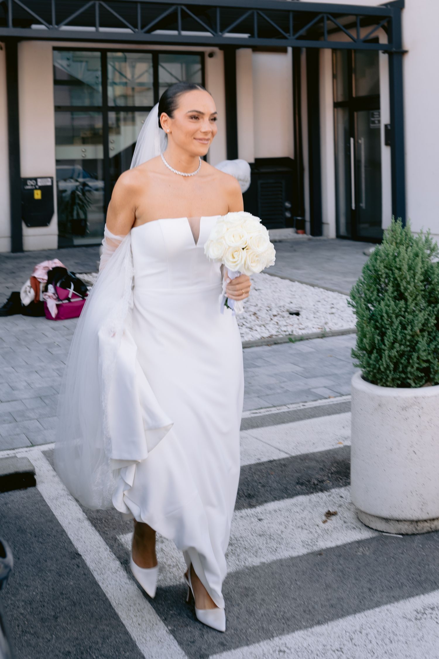 A bride in an elegant off-shoulder white wedding gown carries a white bouquet while walking outside a building.