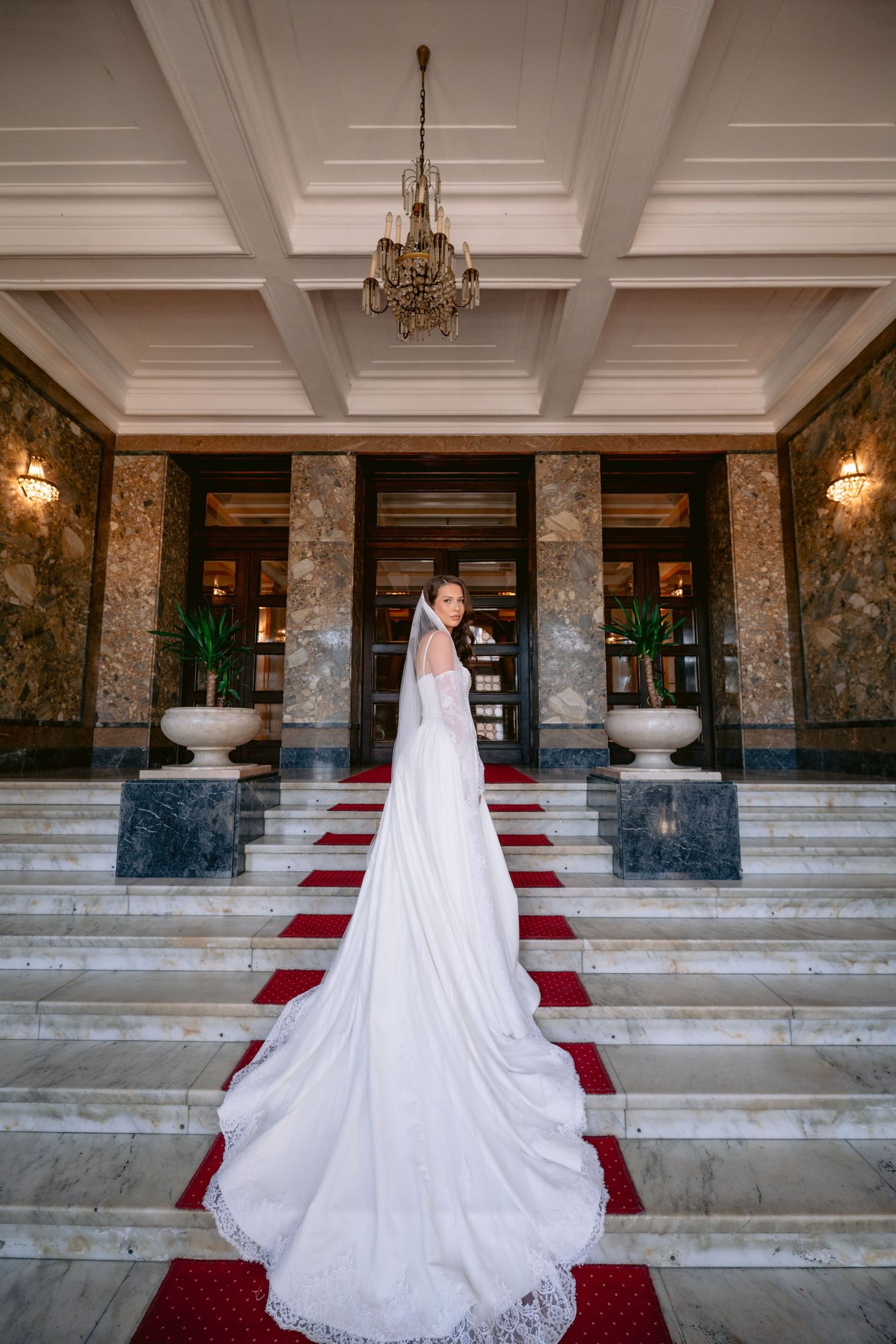 Elegantna bijela vjenčanica sa dugačkim šlepom izložena na veličanstvenom mermernom stepeništu sa crvenim tepihom i lusterom u Banskom Dvoru