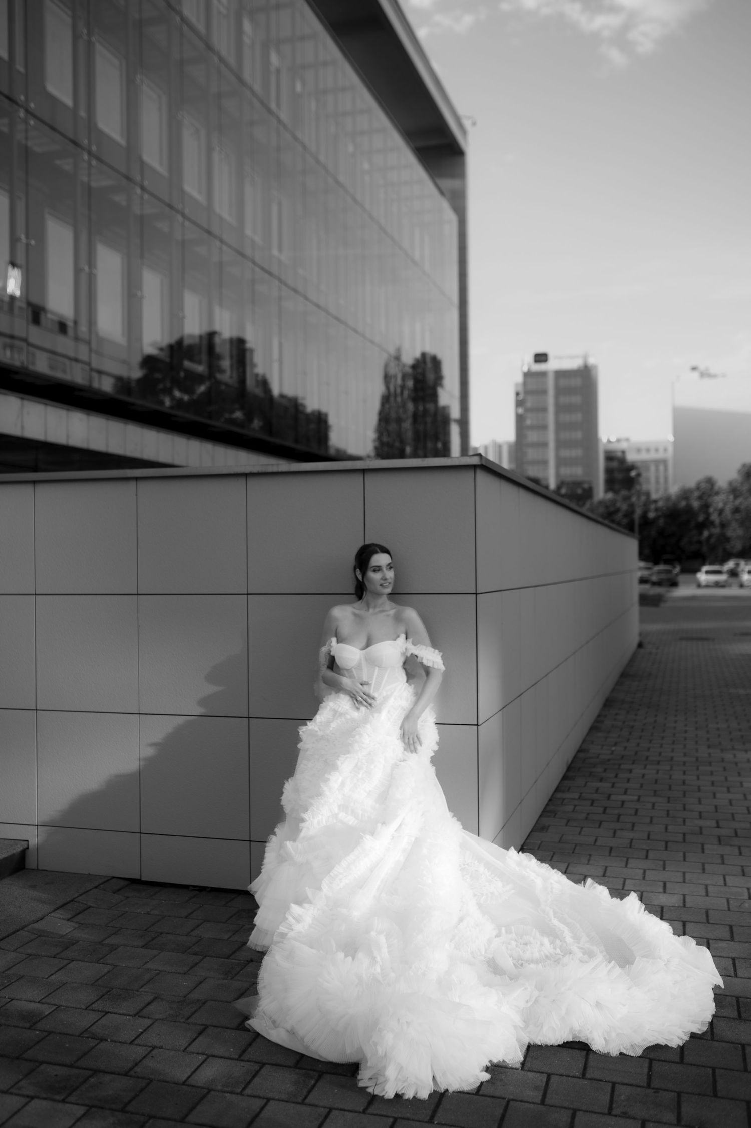 Bride in elegant white ruffled ball gown poses against modern glass building in black and white urban wedding photo.