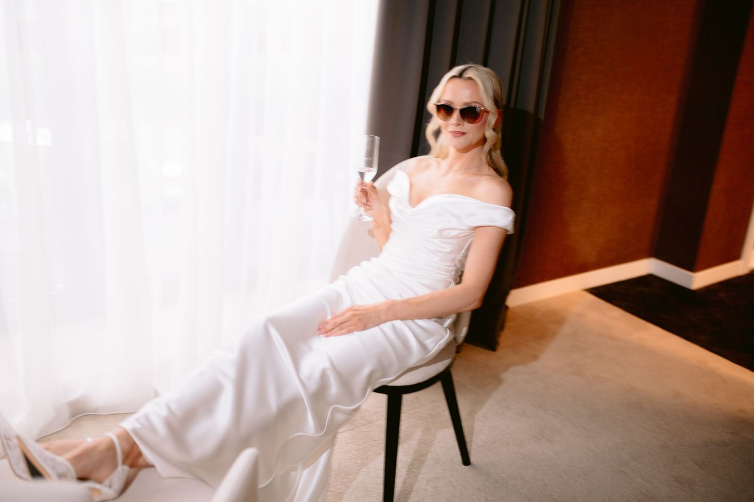 Blonde woman in white off-shoulder jumpsuit and sunglasses holding champagne glass while relaxing on chair near window.