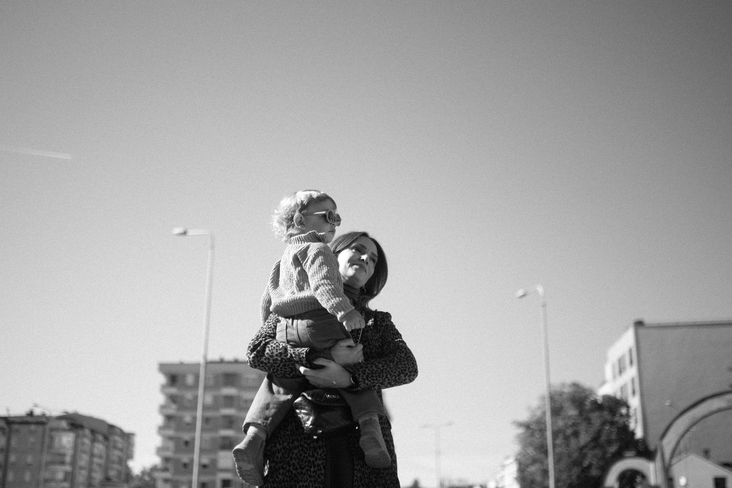 Black and white photo of a woman in a hat holding a small child in her arms outdoors, with city buildings and street lamps behind them.