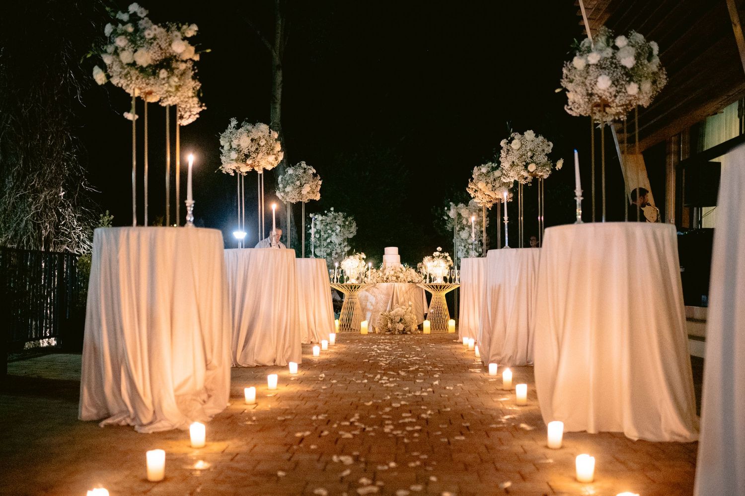 Elegant outdoor wedding aisle lit by candles with white draped pedestals and floral arrangements at night.