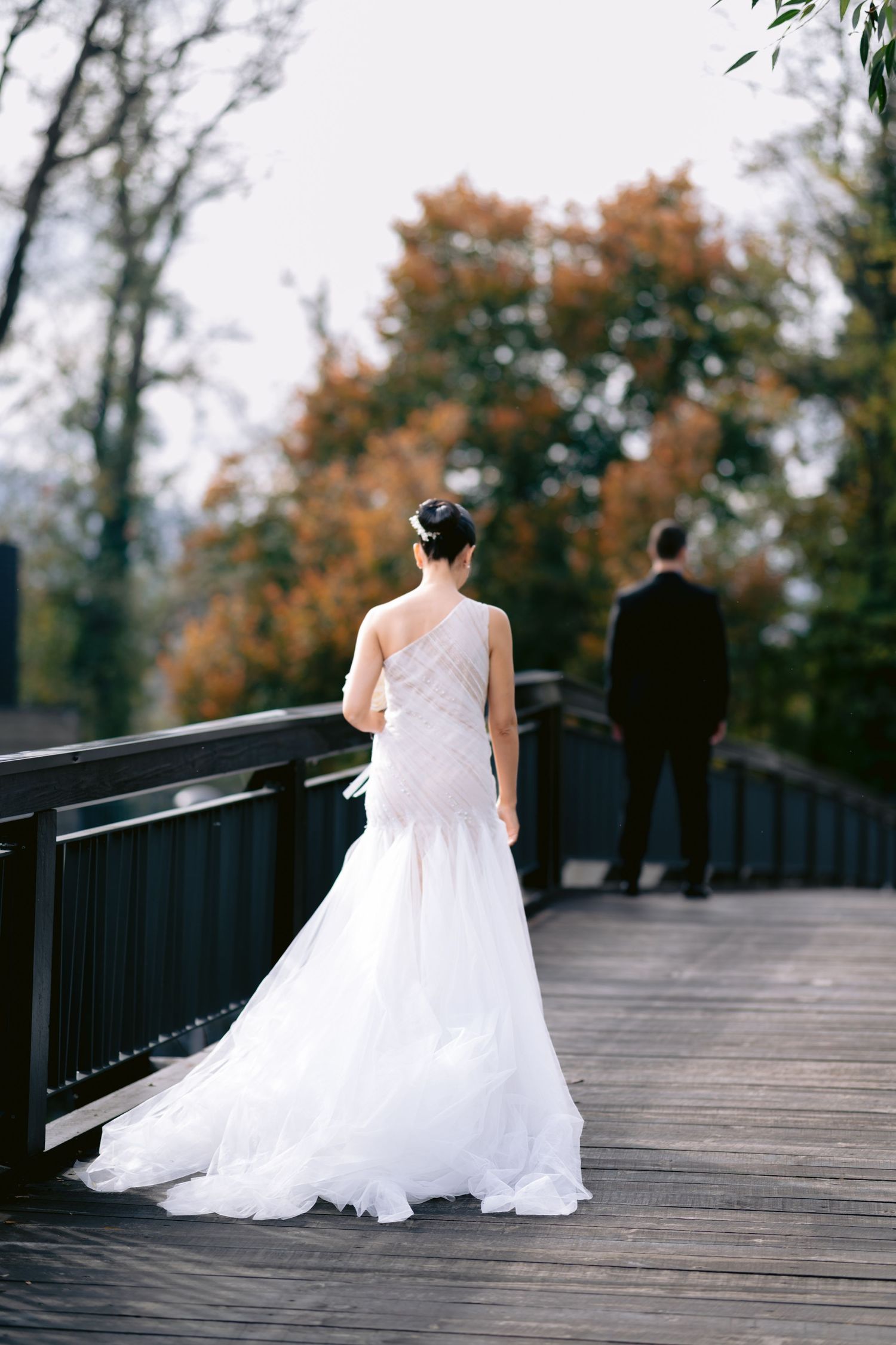 Bride in white gown and groom in black suit on wooden bridge surrounded by autumn trees.