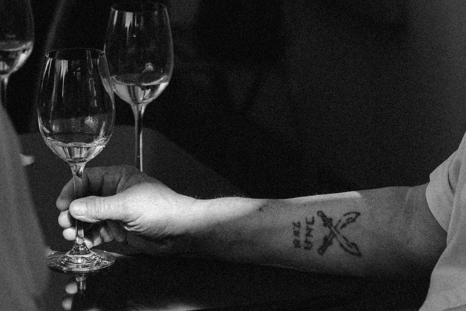 Crno-bijela fotografija tetovirane podlaktice oslonjene na šank pored elegantnih čaša za vino.