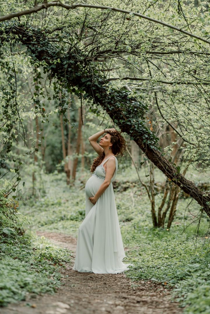 Pregnant woman in white dress standing gracefully in lush green forest under ivy-covered tree branch.