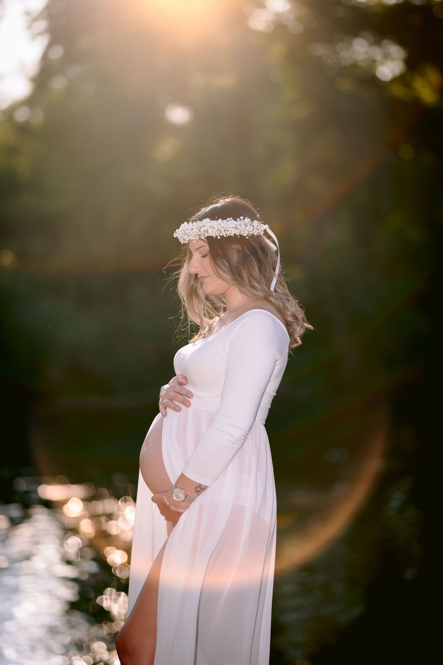 Pregnant woman in white dress and floral crown holds baby bump in golden sunset light near a reflective pond.