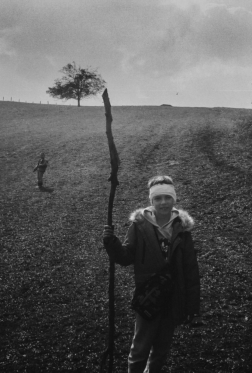 A black and white photograph shows someone in a winter coat holding a tall walking stick on a hillside with a lone tree.