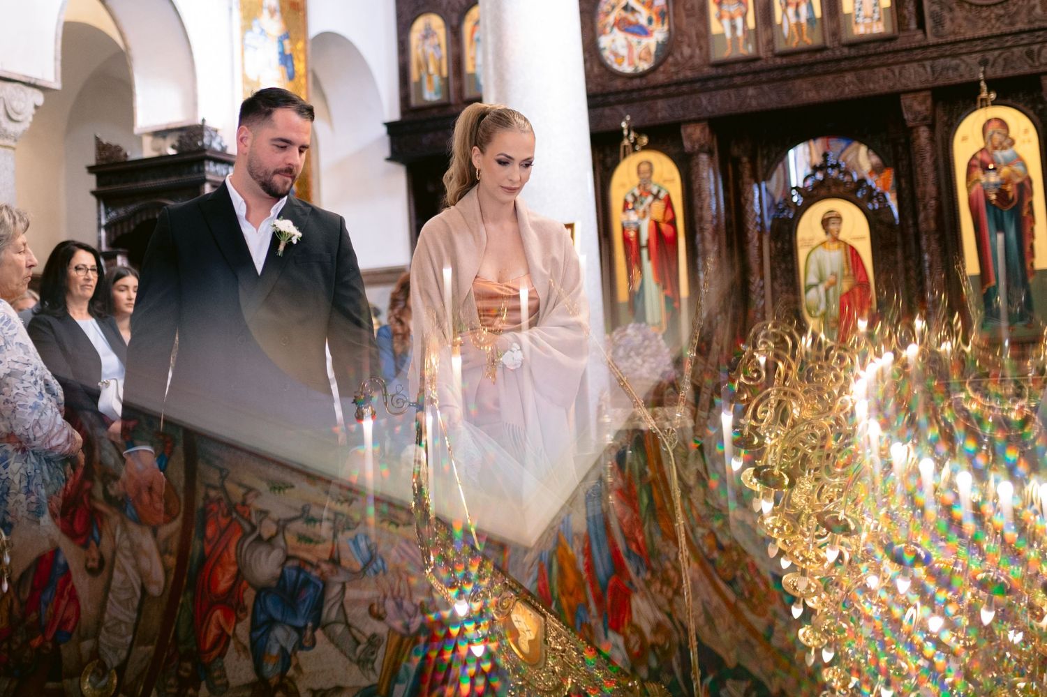 A row of lit candles glows warmly inside an ornate Orthodox church with religious icons on display.