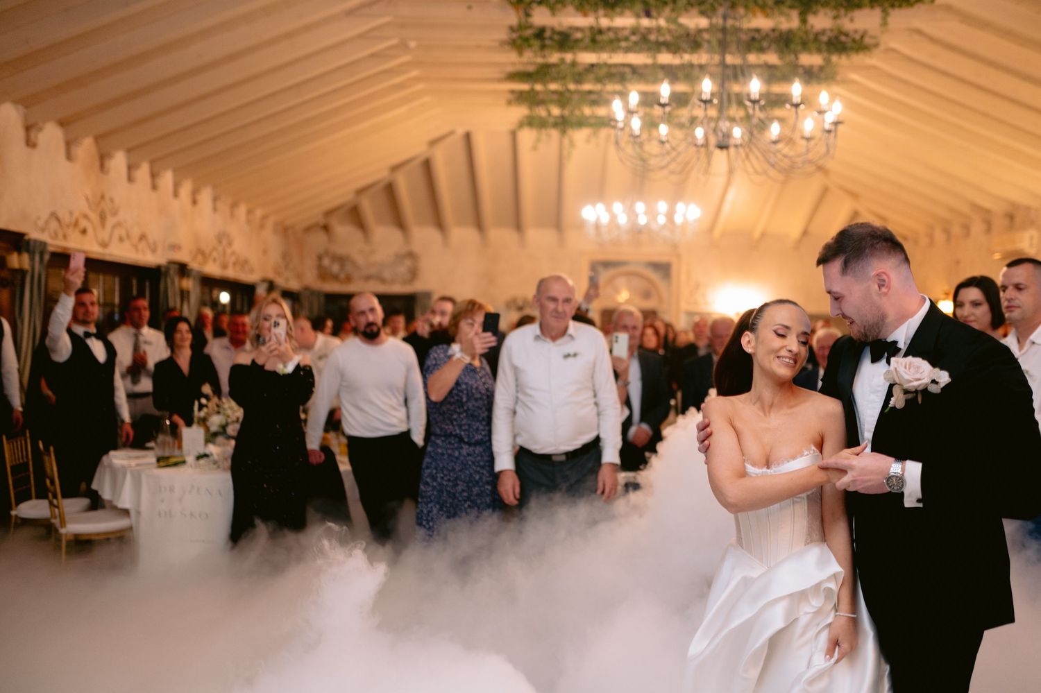 Wedding guests dance and celebrate on the dance floor inside an elegant white tent reception venue with chandeliers.