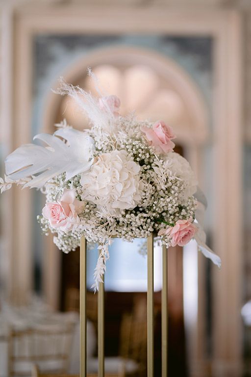 Elegant wedding centerpiece with white peonies, pink roses and baby's breath on gold stand against blurred background.