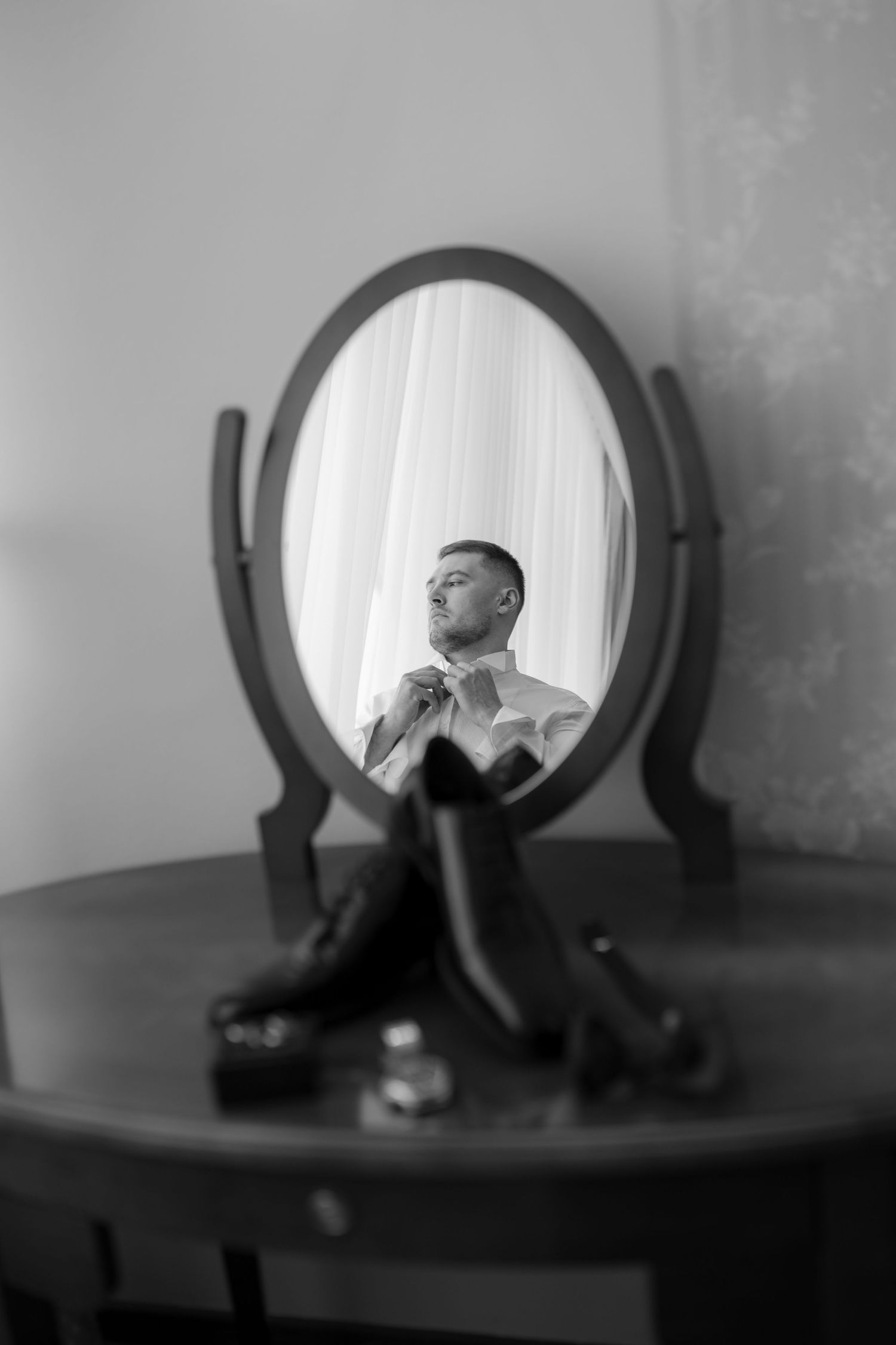 Black and white reflection in oval vanity mirror shows person adjusting tie while getting ready.