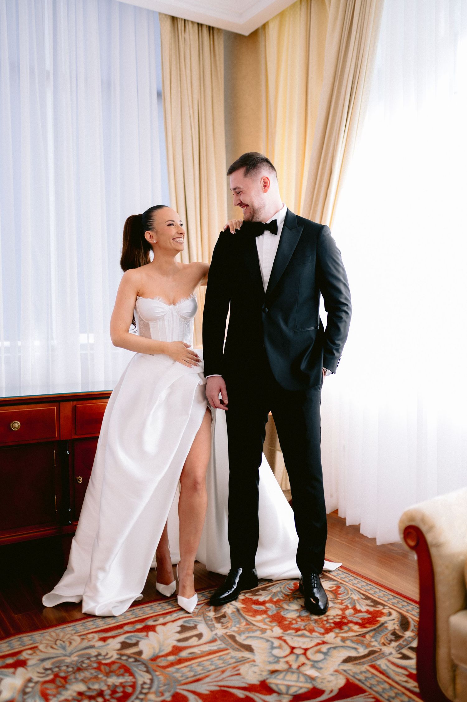 A couple in formal wedding attire shares playful moments in a hotel room with elegant decor and patterned carpet.