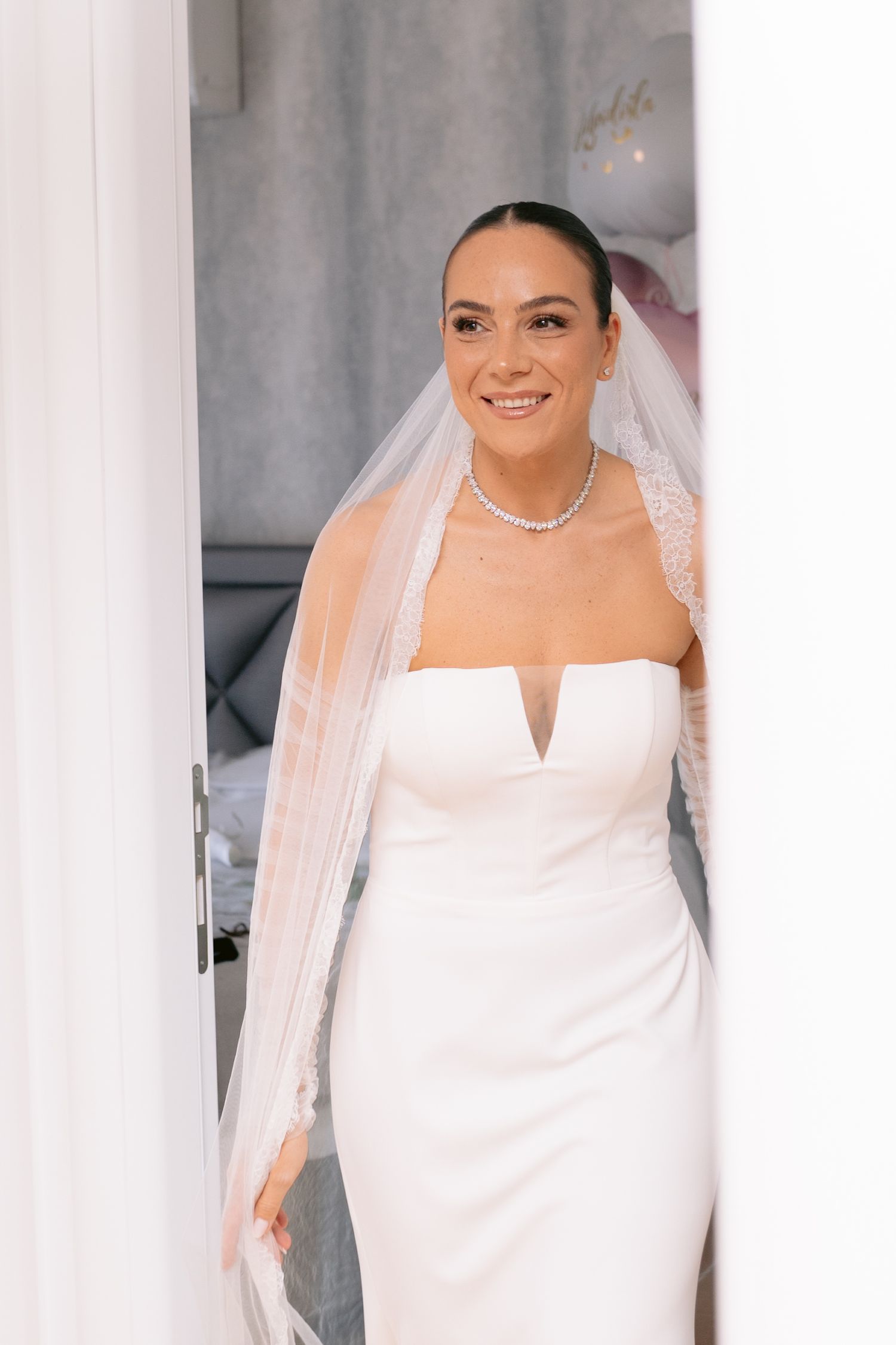 A bride in an elegant white strapless wedding dress and veil smiles while wearing a delicate necklace.