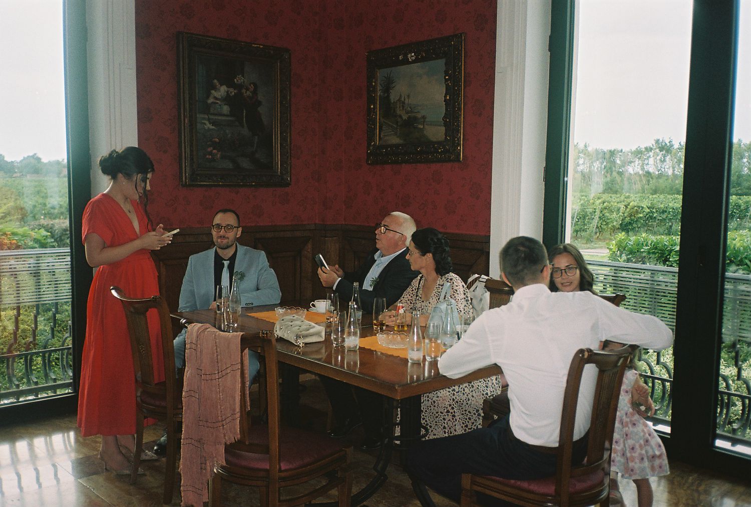 People gathered around a dining table on a balcony with red walls and framed artwork in a vintage setting.