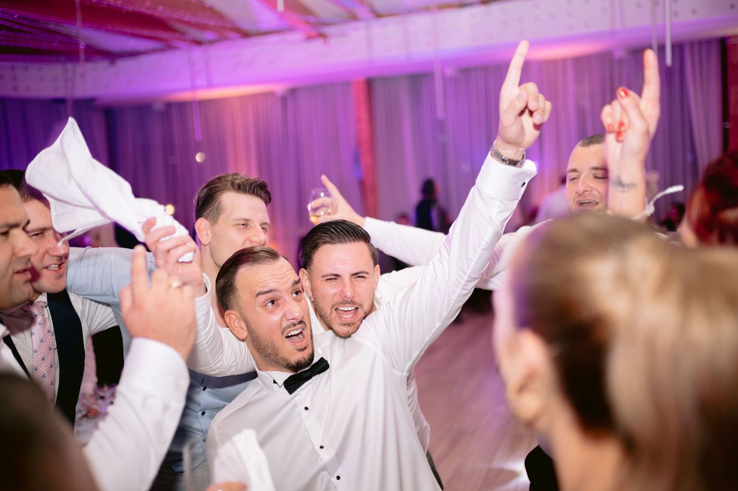 Energetic wedding guests dancing and celebrating with raised arms at a reception with purple lighting.