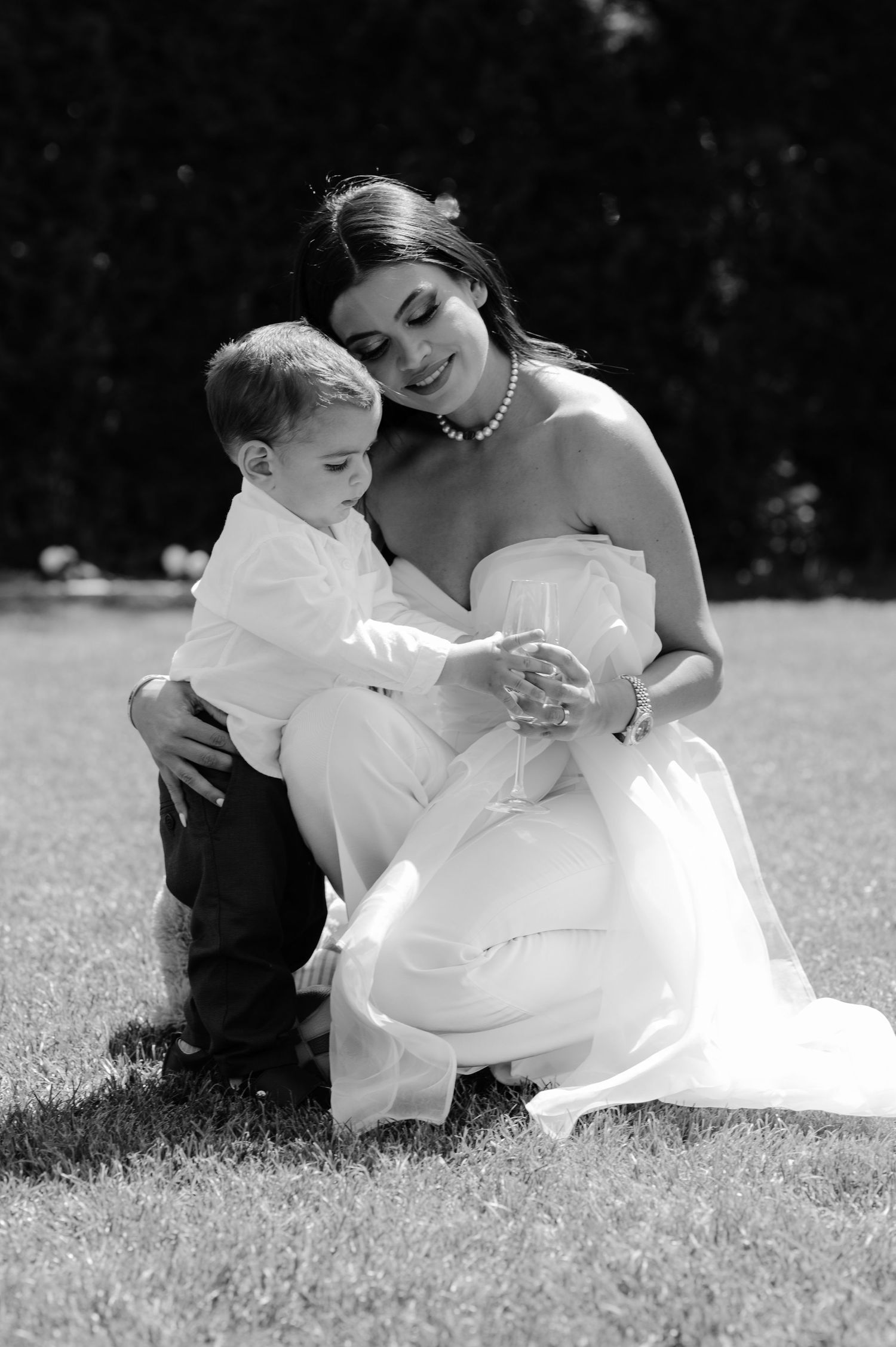 Black and white photo of a smiling bride in a strapless gown kneeling on grass with a young boy in formal attire.