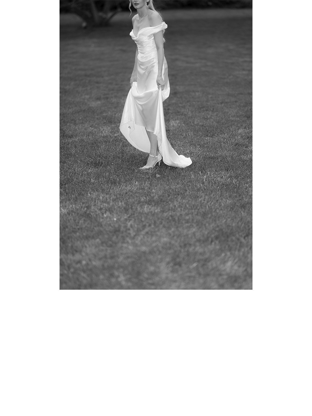 Black and white photo of bride in flowing off-shoulder satin wedding dress walking across green lawn.