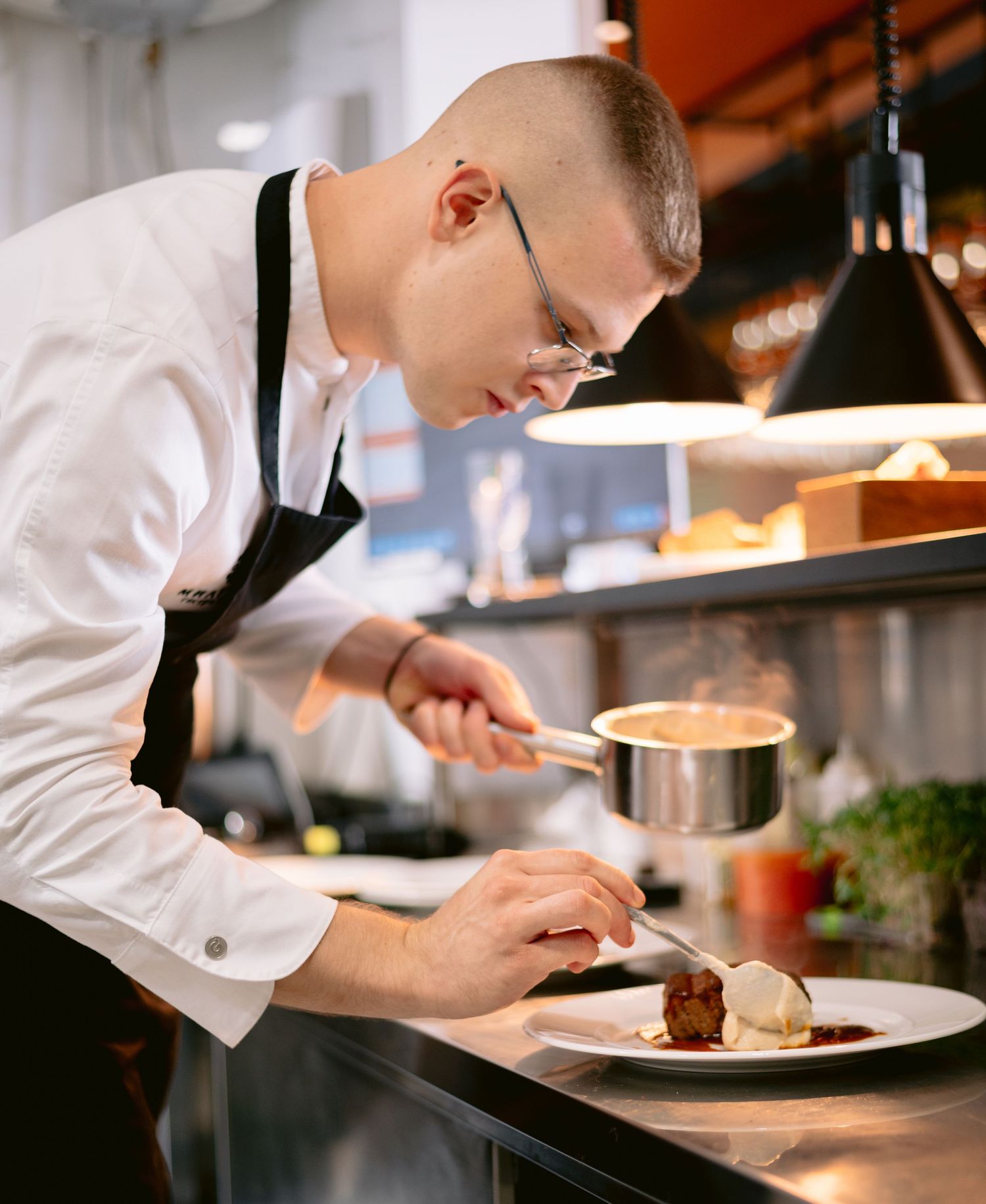A chef in white uniform and glasses carefully plates a gourmet dish using a saucepan in a professional kitchen.