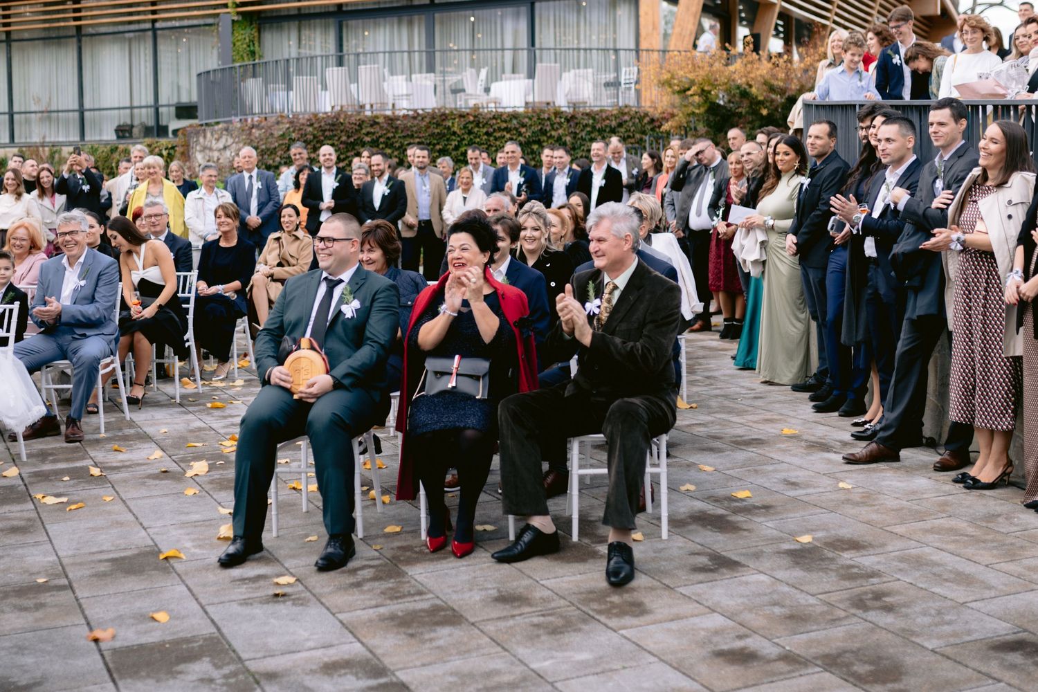 Guests seated and standing applaud at an outdoor wedding ceremony on a paved terrace with autumn foliage.