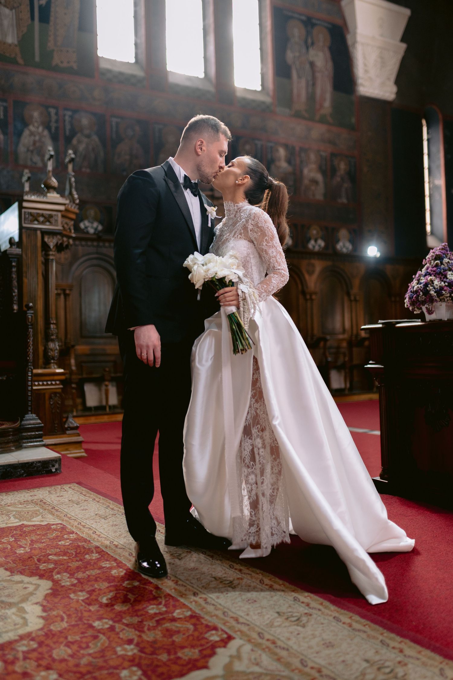 Bride and groom share a kiss at the altar in an ornate church, bride holding white bouquet in lace wedding gown.