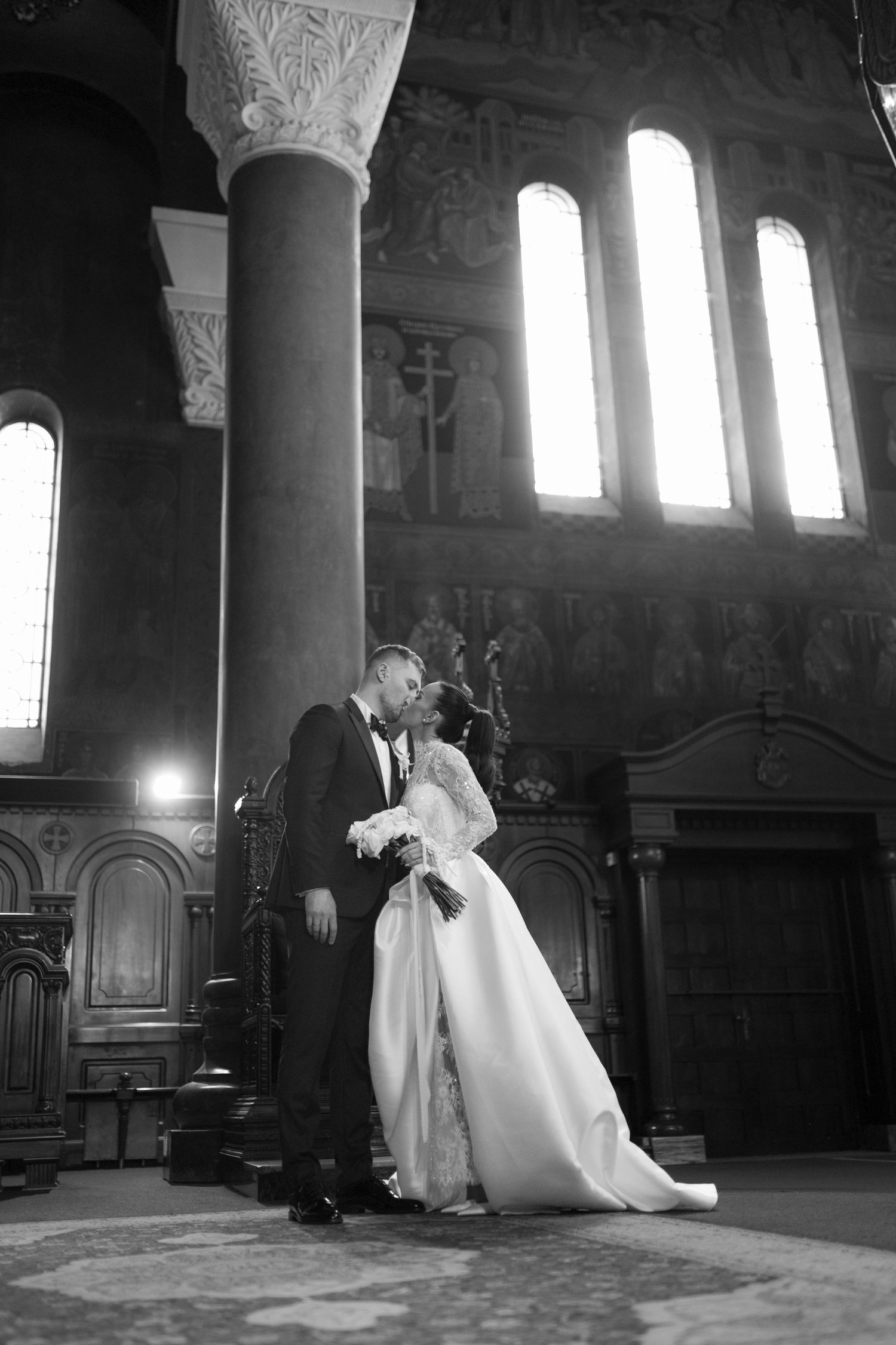 Dramatic black and white photo of wedding couple kissing in church with arched windows.
