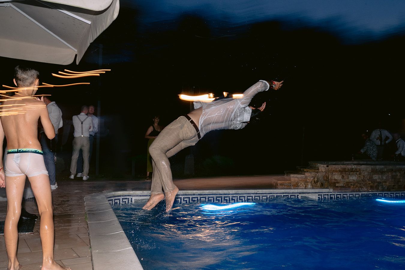 People jumping into an illuminated swimming pool at night creating splashing water effects.