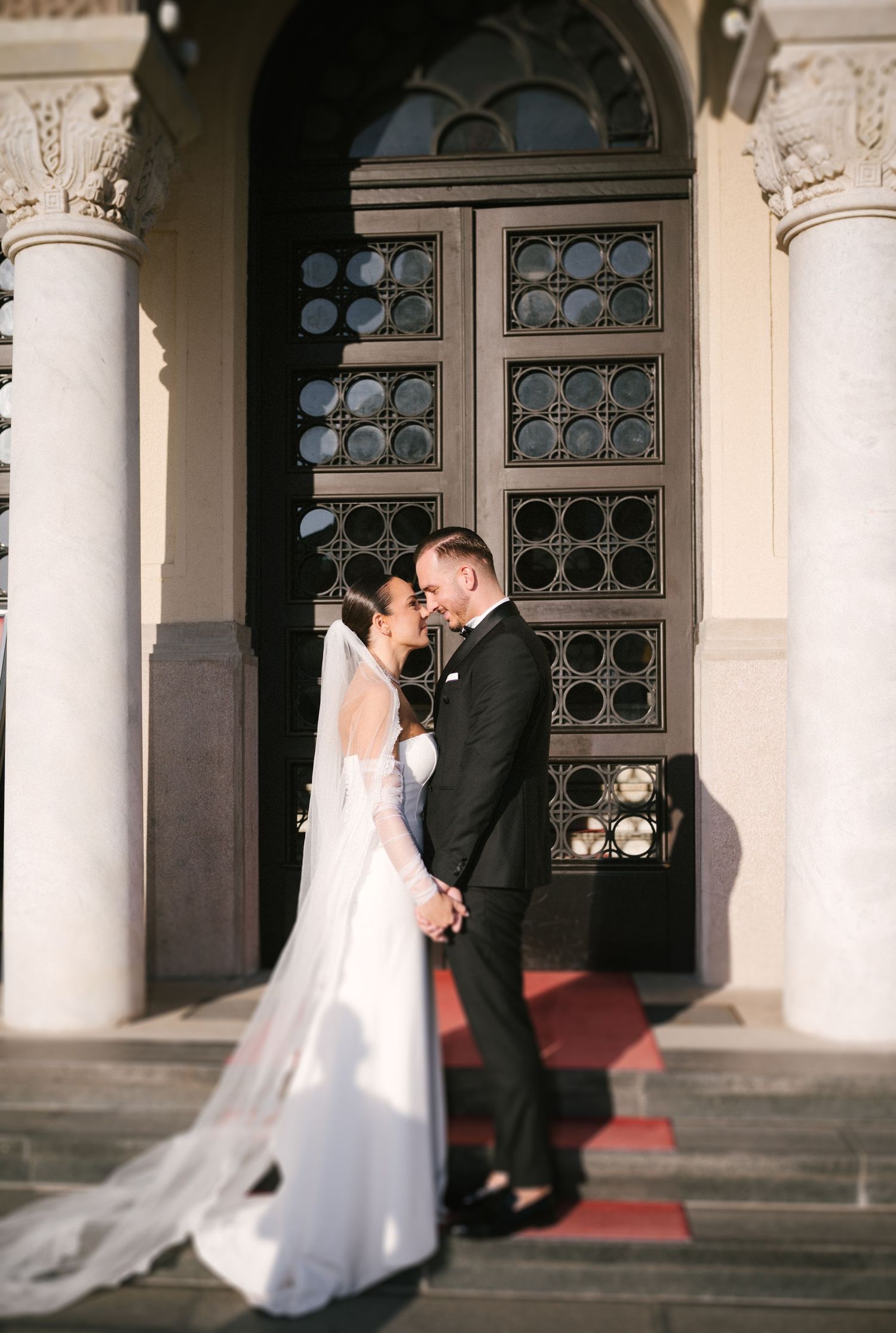 A bride and groom share a romantic kiss under an elegant archway with ornate windows at dusk.