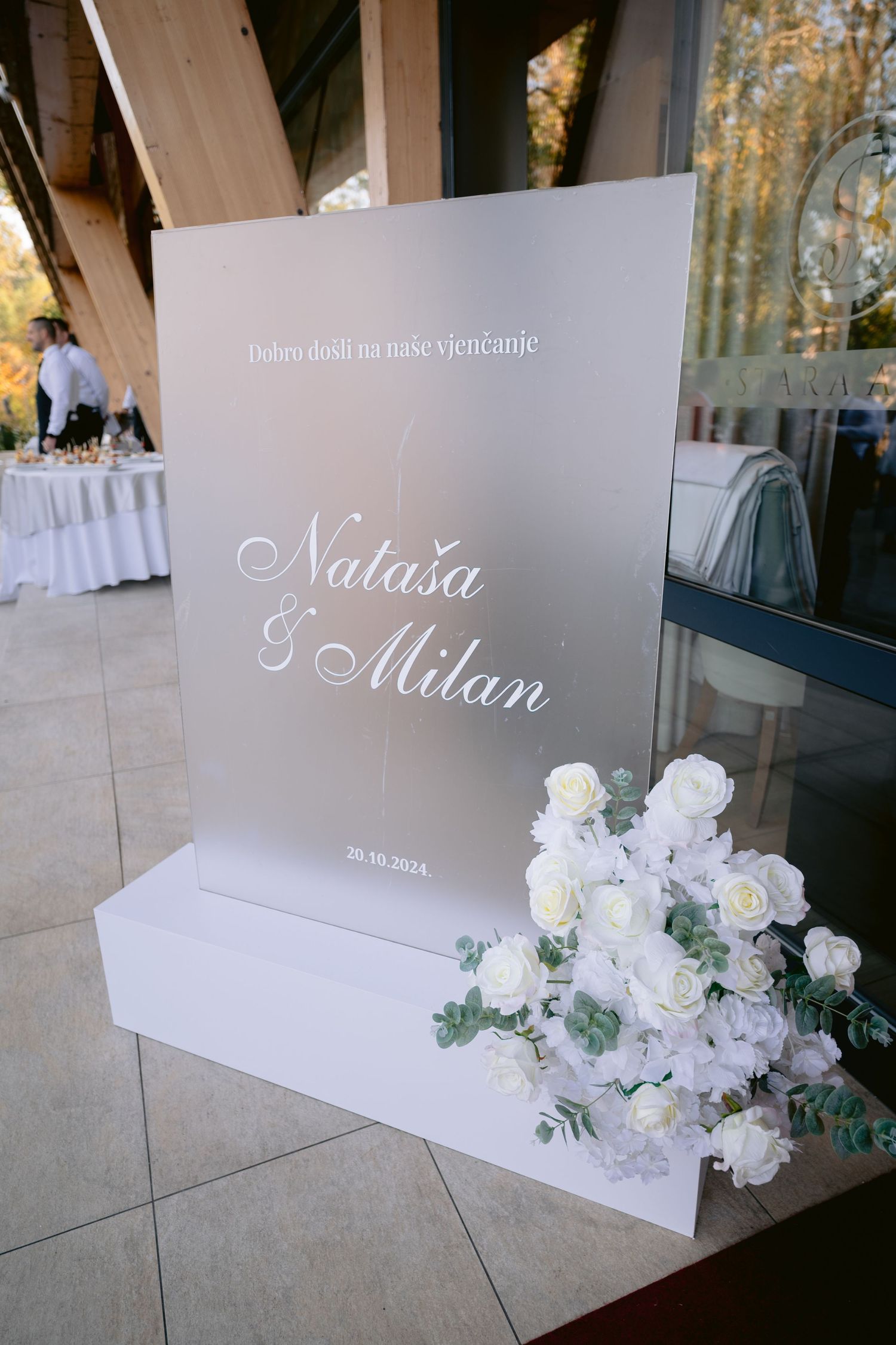 Elegant white wedding signage with floral arrangements displayed on modern white platforms at an outdoor venue.