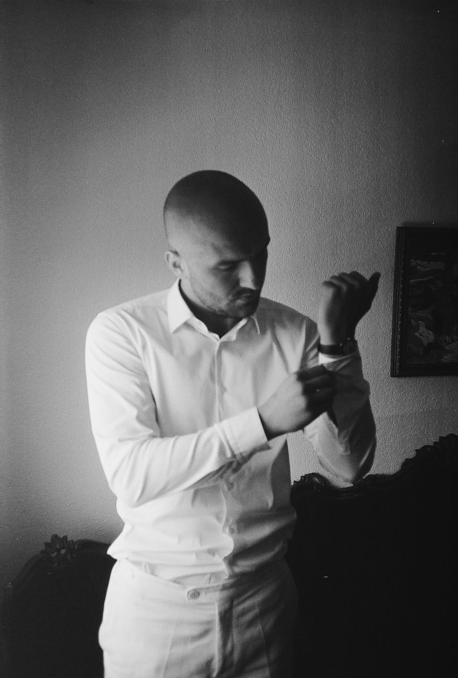 Black and white photo of a bald man in a white dress shirt fastening his cufflinks indoors.