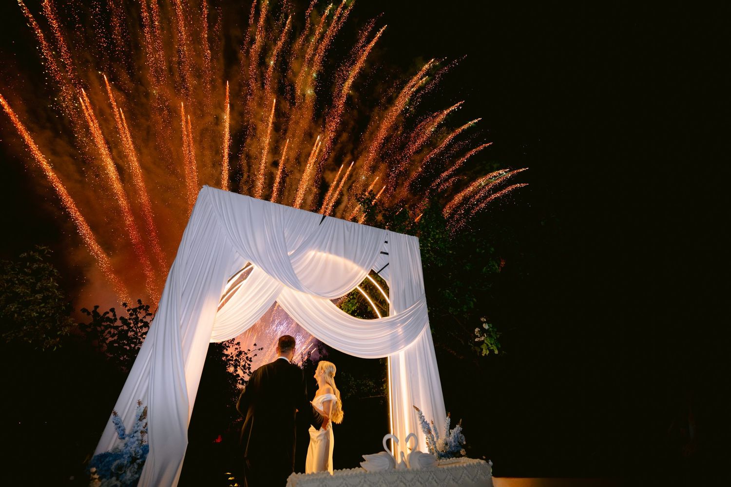 Couple stands under white draped wedding chuppah with stunning fireworks exploding in night sky behind them.