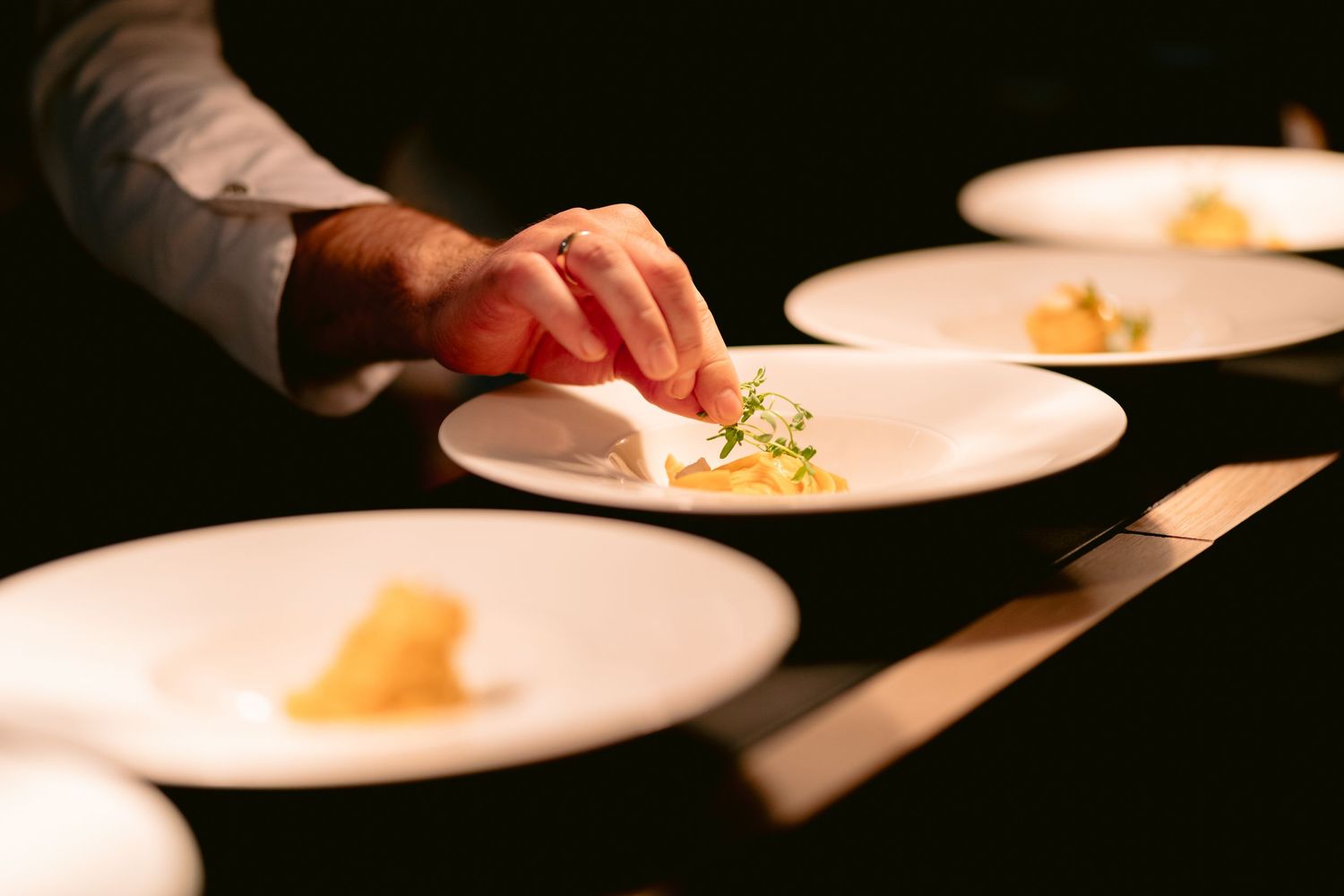 A hand garnishes elegant plated dishes on white plates in a fine dining restaurant setting.
