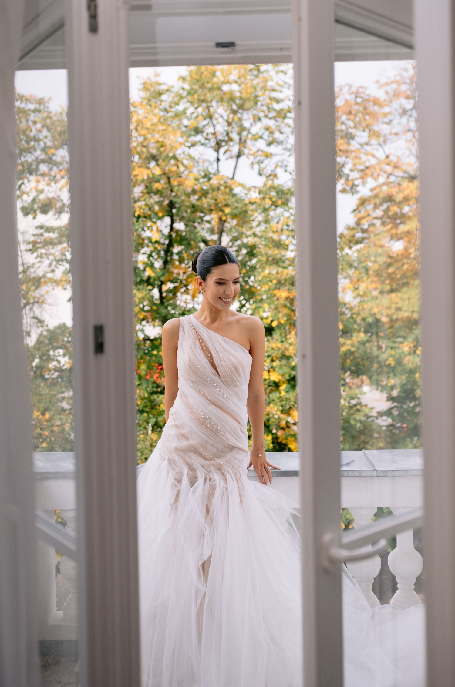 Elegant bride in white gown poses on classic white columned porch with fall foliage in background.
