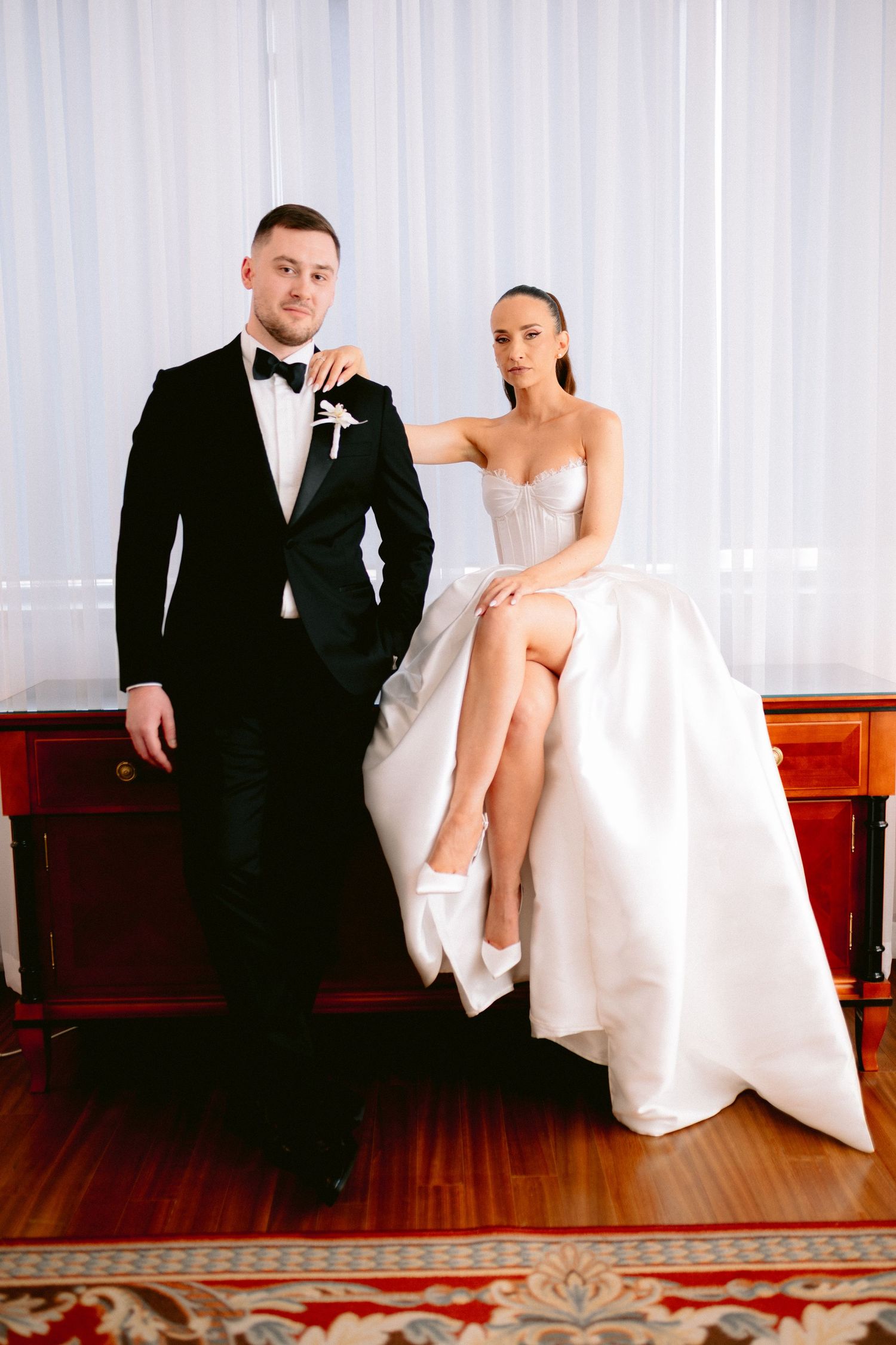 Wedding couple in formal attire posing against white curtains in stylish indoor setting.