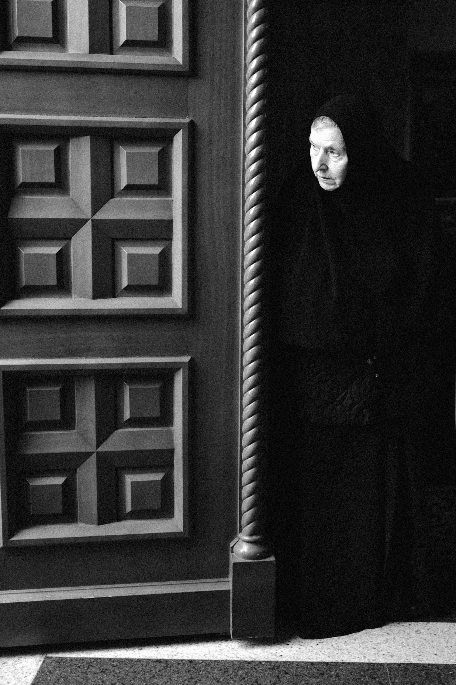 Dramatic black and white portrait through doorway showing figure in dark clothing.
