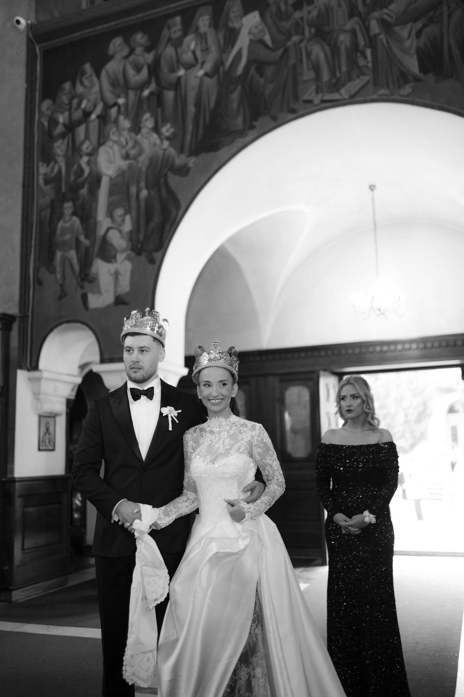 A black and white photograph shows a formal wedding ceremony with people in elegant attire standing in an arched hallway.