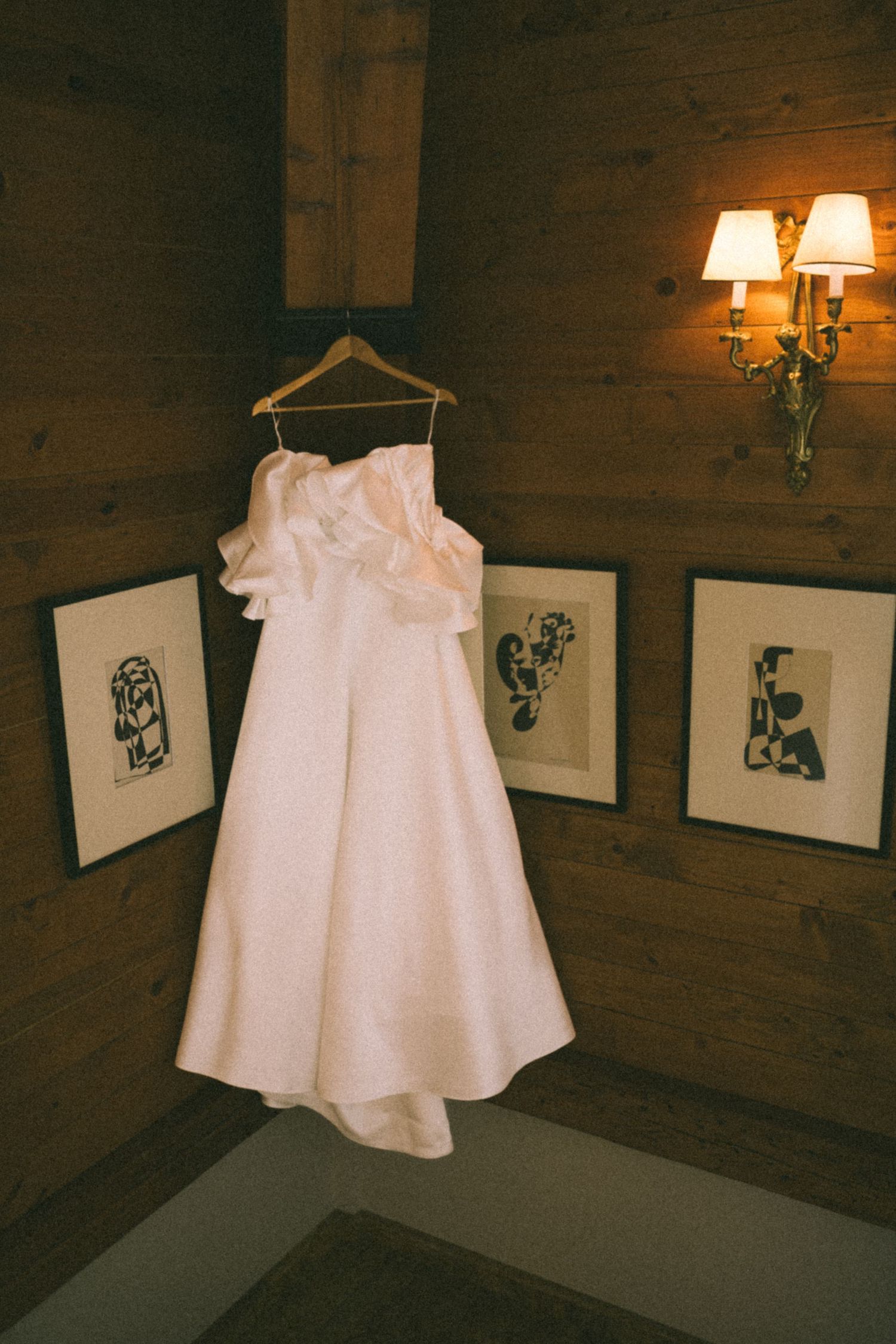 A white wedding dress hangs on a wooden hanger against rustic wood paneling with framed artwork and a wall sconce.