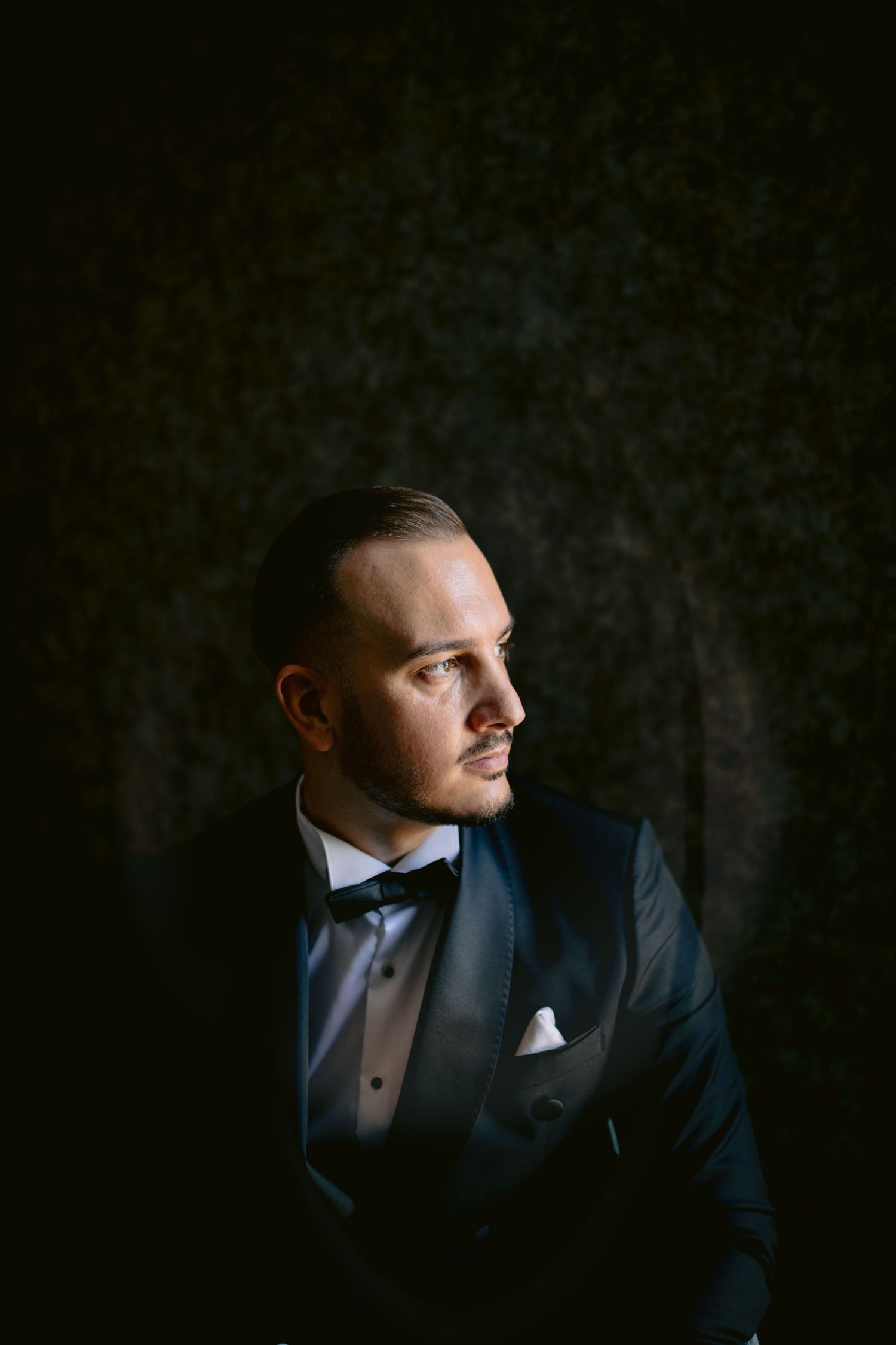Dramatic portrait of a person in formal black tuxedo and bowtie looking sideways against dark background.