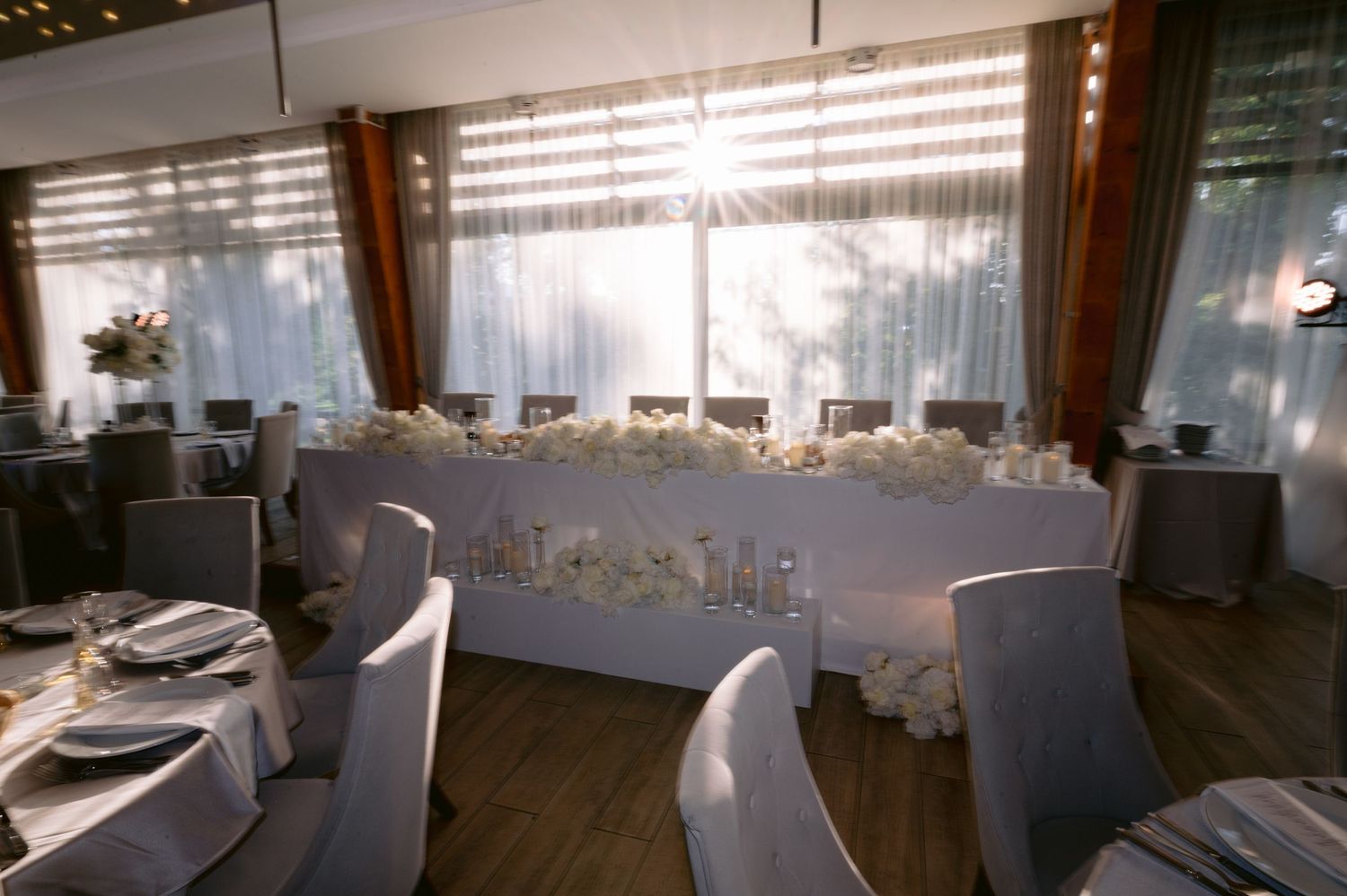 Elegant wedding reception venue with white floral centerpieces, draped windows, and neatly arranged tables with white chair covers.