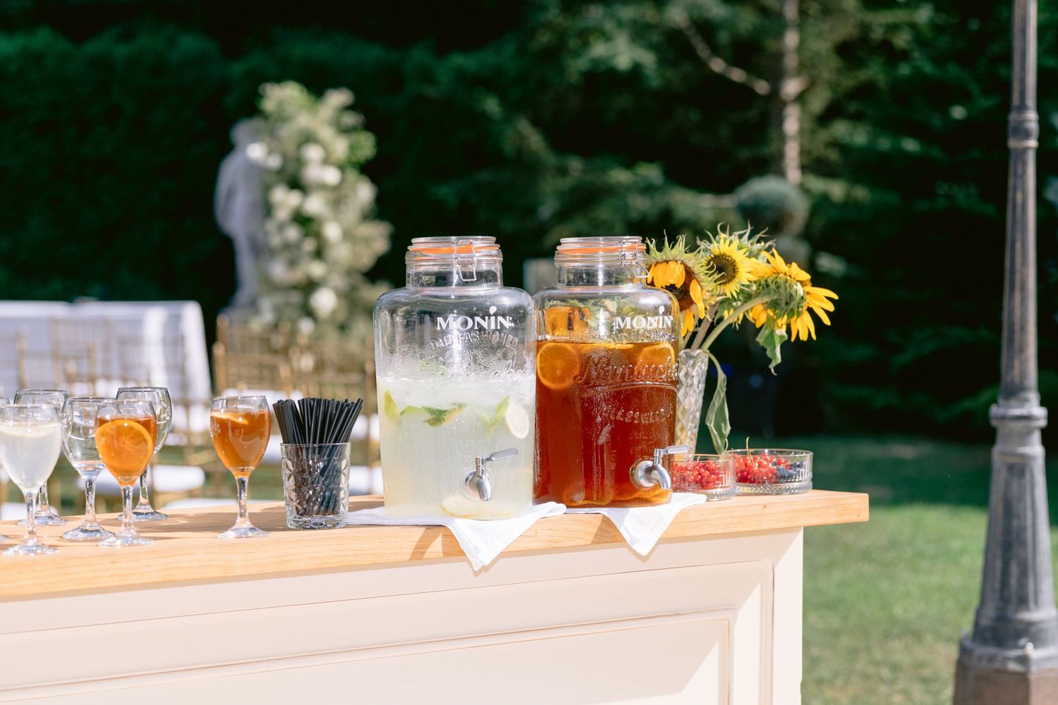 Outdoor drink station with lemonade and iced tea dispensers, sunflowers, and glasses on a white bar table.