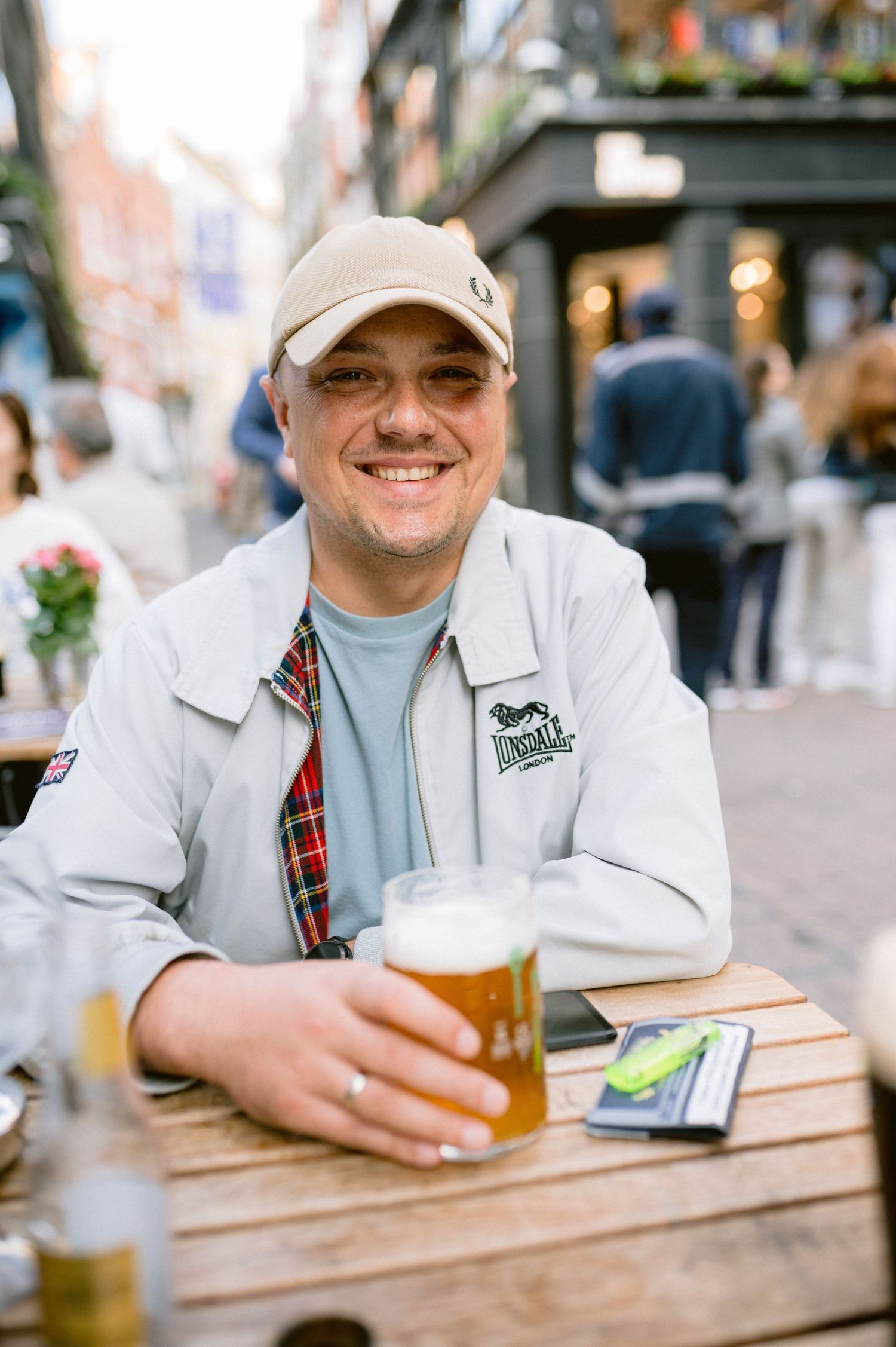 Someone wearing a baseball cap and light jacket sits at an outdoor table with a glass of beer on a sunny day.