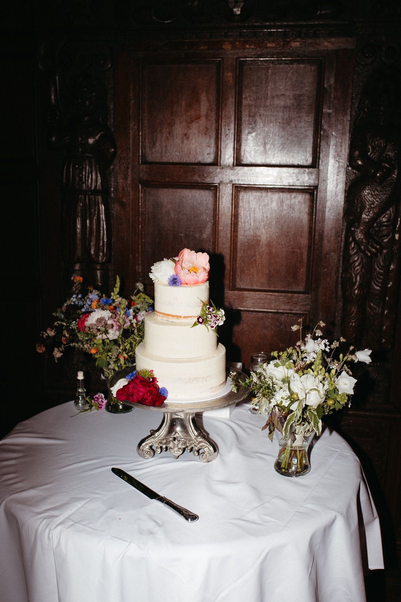 A three-tiered white wedding cake with floral decorations sits on a silver stand next to flower arrangements on a white tablecloth.
