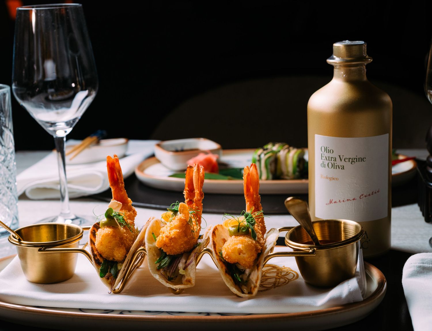Elegant shrimp tacos served in gold holders with dipping sauces and olive oil bottle on white plate.