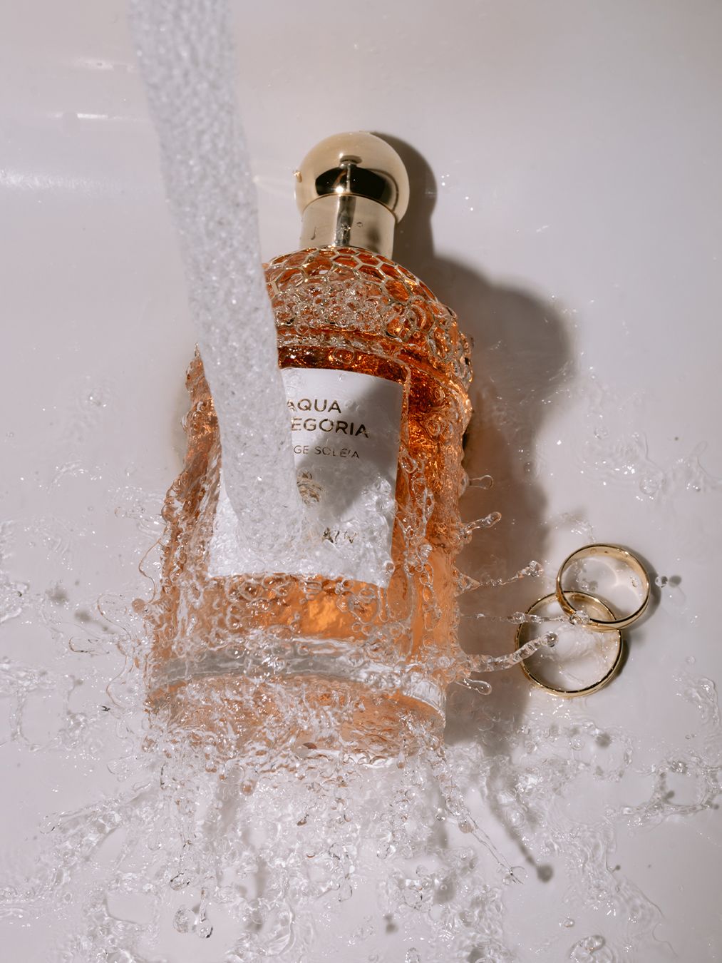 Aqua Allegoria perfume bottle submerged in splashing water with gold ring accent on white surface.