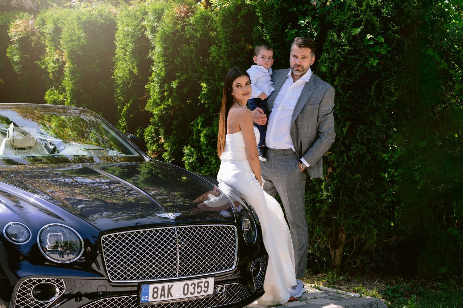 Elegant family posing beside a luxury Bentley car in a lush green garden setting, dressed in formal wedding attire.