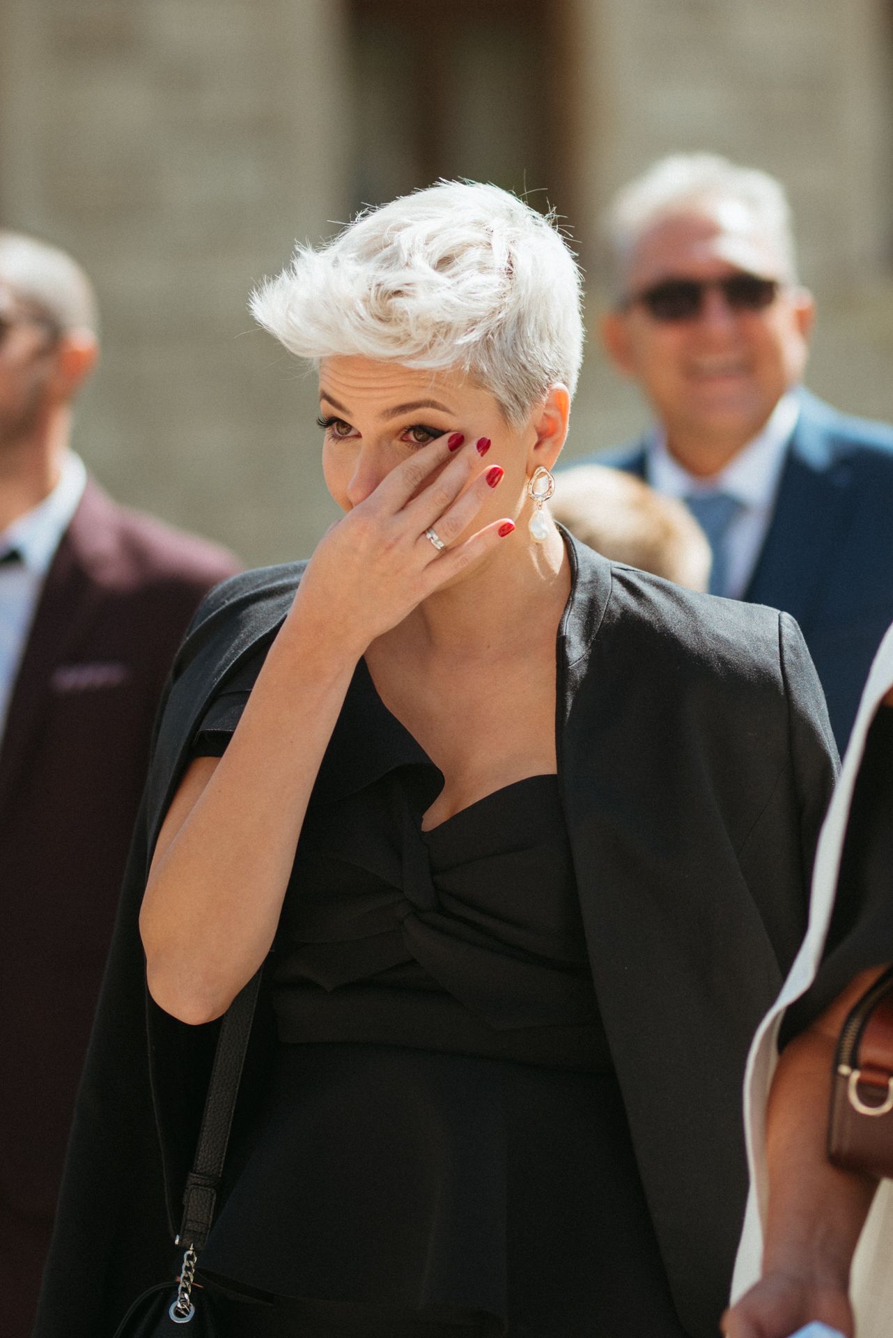 Woman with short white hair in black outfit covering her face with red-nailed hand outdoors.