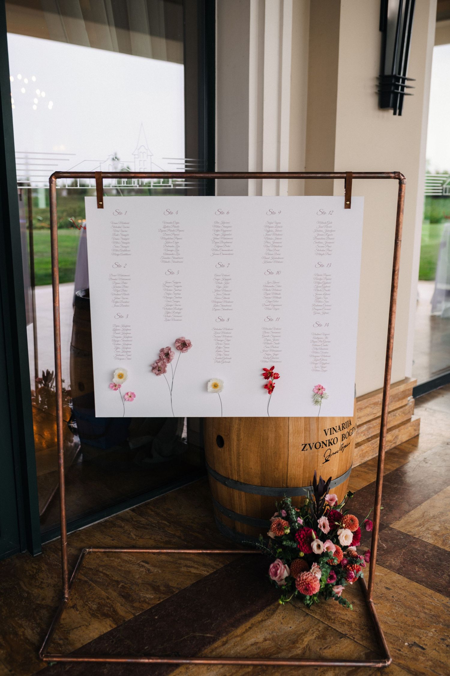 A wedding seating chart display decorated with pink and red flowers on a wooden barrel at a rustic venue.