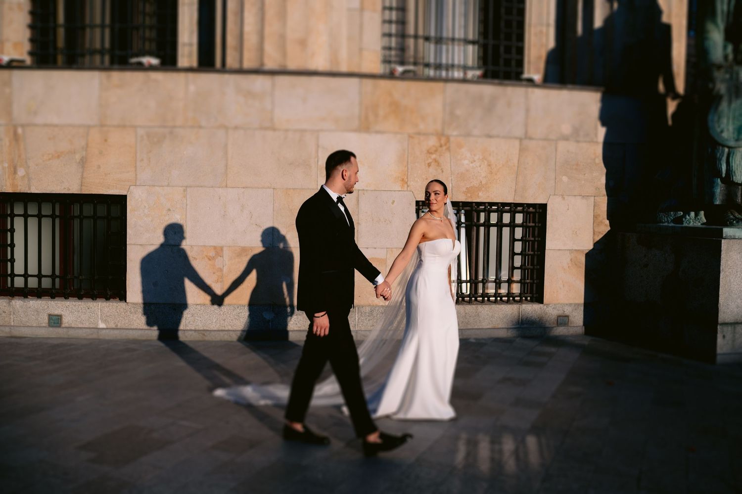 A couple in wedding attire walks together while their shadow creates a romantic silhouette on a stone wall.