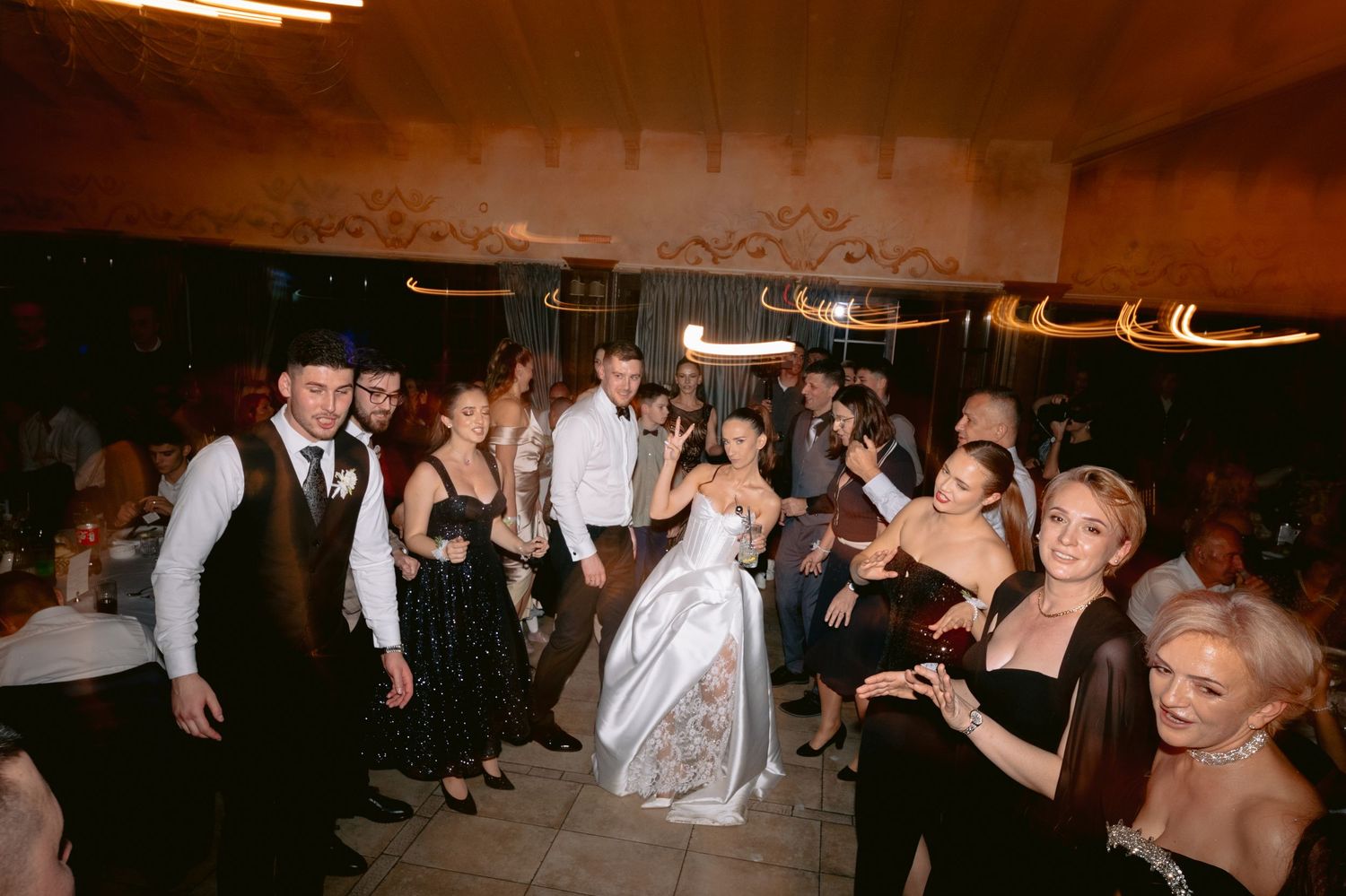 A group of wedding guests dance together on a dimly lit dance floor during an evening reception celebration.