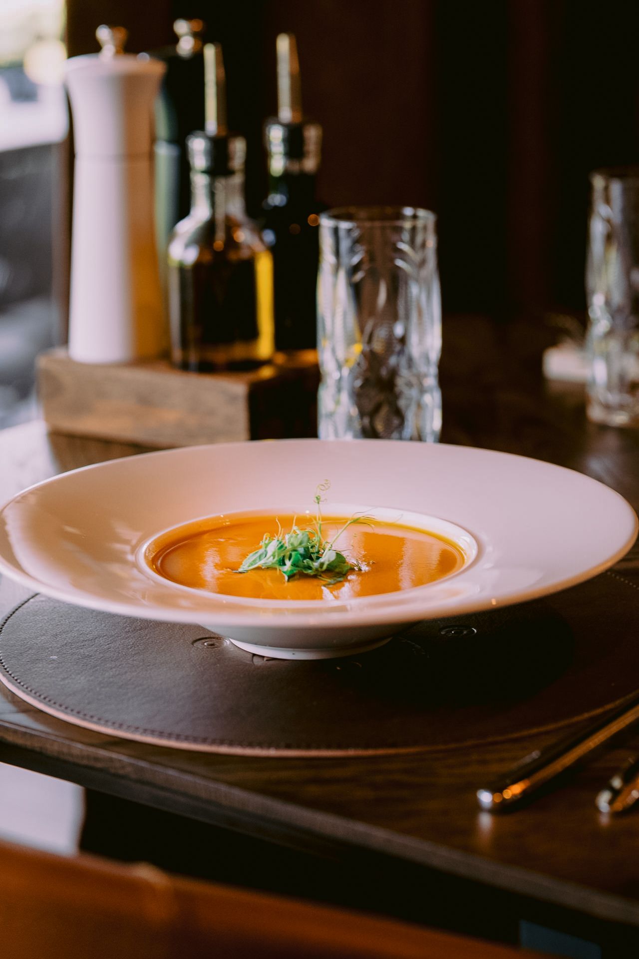 Orange soup garnished with microgreens served in an elegant white bowl on wooden table.
