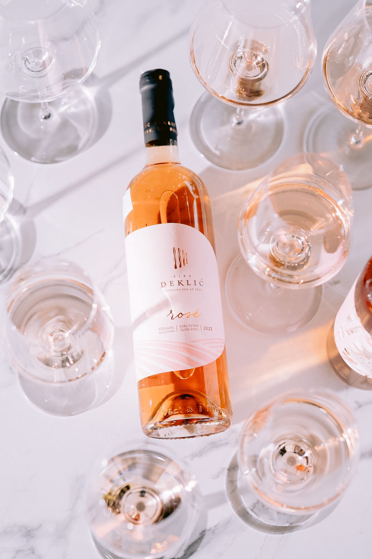 Rose wine bottle surrounded by empty wine glasses on marble background.